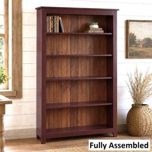 May include: A tall, dark red wooden bookcase featuring five open shelves and a vertical plank back panel in a natural brown wood finish. The unit includes a top shelf with a small potted plant and several books. The text Fully Assembled is visible.