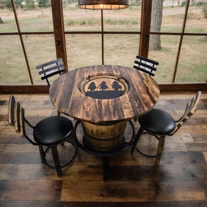 May include: A rustic wooden table with a decorative metal insert, supported by a barrel base. Four black leather-seated chairs with wooden backs surround the table. The table and chairs are set in a room with large windows.