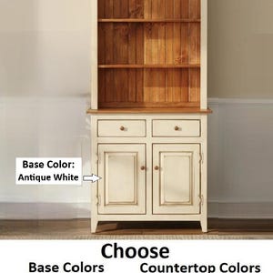 May include: A hutch with an antique white base and a walnut-colored countertop and backing. The unit features two upper shelves, two drawers with small knobs, and two lower cabinet doors. Text on the image lists available base and countertop colors.