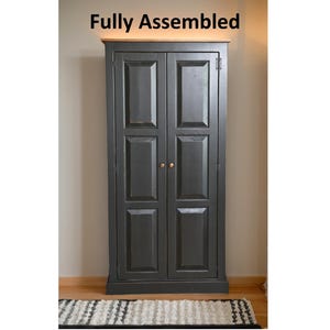 May include: A tall, dark gray cabinet with two doors, each featuring three rectangular recessed panels. The cabinet has a natural wood-toned top surface and two small, round metallic knobs. The text "Fully Assembled" appears above the cabinet.