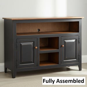 May include: A black media console with a natural wood-toned top and interior shelving. The unit features two side cabinets with single doors and a central section with two open shelves. The text "Fully Assembled" is printed in the bottom corner.