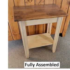 May include: A small wooden side table with a two-tone finish. The tabletop is stained a dark brown, while the legs and shelf are painted a cream color. The table has a rectangular top and a lower shelf for storage. The text "Fully Assembled" is at the bottom.