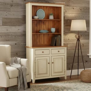 May include: A tall wooden hutch featuring a cream-colored frame and a natural wood-toned interior back panel. The unit includes three open shelves holding ceramic plates and bowls, two drawers with round knobs, and two lower cabinet doors.