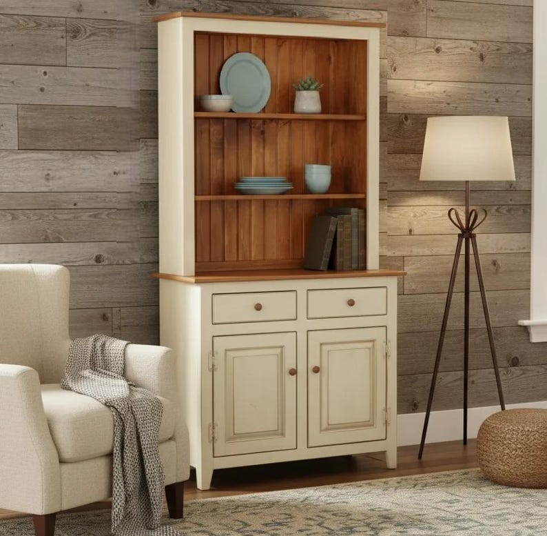 May include: A tall wooden hutch featuring a cream-colored frame and a natural wood-toned interior back panel. The unit includes three open shelves holding ceramic plates and bowls, two drawers with round knobs, and two lower cabinet doors.