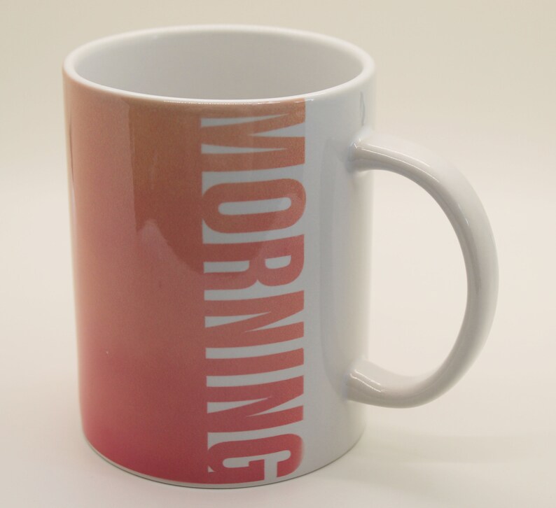 K&ouml;nnte beinhalten: Wei&szlig;e Keramiktasse mit einem Ombre-Design von Rosa zu Orange und dem Wort "MORNING" in Wei&szlig; auf der Seite.