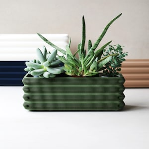 Op de afbeelding: Een donkergroene rechthoekige plantenbak met een gegolfd ontwerp, gevuld met diverse vetplanten en kleine planten. De plantenbak heeft een textuuroppervlak. Andere plantenbakken in wit, marineblauw en bruin zijn op de achtergrond.