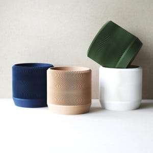 May include: Four cylindrical planters in navy blue, beige, white, and green. Each planter has a textured, ribbed exterior and a solid-colored base. The planters are arranged in a group, with one tilted on top of another.