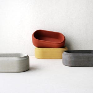 Könnte beinhalten: Drei ovale Beton-Pflanzkübel in verschiedenen Farben: Grau, Gelb und Rot. Die Kübel sind übereinander gestapelt.