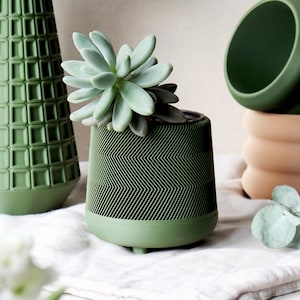 May include: A green ceramic planter with a chevron pattern, holding a succulent plant. The planter is on a white cloth, with other green and peach-colored ceramic vases in the background. The succulent has pale green leaves.