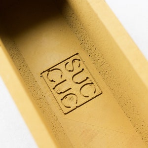 May include: A close-up of a yellow planter with the word "SUC" embossed on the inside.