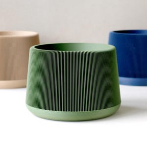 Op de afbeelding: Drie moderne, geribbelde plantenbakken in minimalistische stijl. De voorste plantenbak is een gedempt groen, met een afgeronde basis en een brede opening. Een bruine en een blauwe plantenbak staan op de achtergrond. De plantenbakken zijn waarschijnlijk van keramiek.