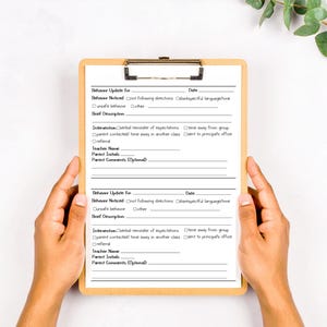 May include: A light brown wooden clipboard holding a white behaviour update form. The printed document features two identical sections with fields for date, behaviour noticed, brief description, intervention, teacher name, parent initials, and comments.