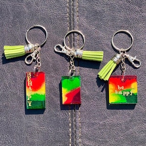 May include: Three colourful resin keychain charms with green, red and yellow accents. The charms are attached to silver key rings with green tassels. The charms feature the words "be happy" and "be happy" in gold lettering.