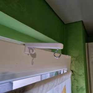 Könnte beinhalten: Weißer Kunststoff-Vorhangstangenhalter mit einem Metallhaken, der an einem Fensterrahmen befestigt ist.