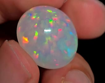 May include: A polished, oval-shaped opal gemstone. The opal is primarily white with iridescent flashes of green, pink, yellow, and blue throughout its surface. The gemstone is held between two fingers against a dark background.