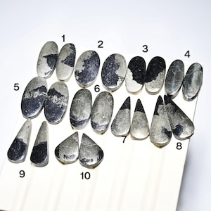 Pode incluir: Uma coleção de dez pedras preciosas de pirita em várias formas. As pedras são principalmente prateadas e pretas, com um brilho metálico. As formas incluem ovais, lágrimas e formas irregulares, dispostas numa superfície branca.