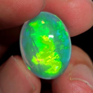 Puede incluir: Una vibrante piedra preciosa de ópalo de forma ovalada. El ópalo muestra un fascinante juego de colores, con destellos vívidos de verde y amarillo sobre un fondo translúcido y azulado. La gema se sostiene entre los dedos, mostrando su superficie pulida.