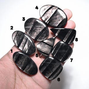 Puede incluir: Ocho piedras de obsidiana plateada pulidas numeradas del uno al ocho. Cada piedra presenta una superficie lisa y reflectante con bandas negras y plateadas metálicas. Las piedras varían en formas ovaladas, triangulares y redondeadas.