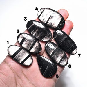 May include: Seven polished oval and teardrop-shaped black obsidian stones with silver sheen inclusions, numbered one through seven. Each stone displays a reflective metallic luster and dark, smooth surfaces with distinct linear patterns.