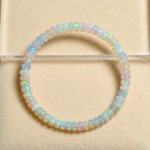Op de afbeelding: Een cirkelvormige armband bestaande uit kleine, doorschijnende, schijfvormige opaal kralen. Elke kraal heeft een melkachtige witte basis met iriserende flitsen van blauw, groen en roze licht. De kralen zijn geregen in een continue lus.