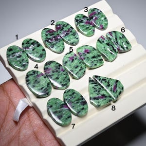 May include: A collection of eight pairs of polished zoisite gemstones with ruby inclusions. The stones display a mottled green pattern with streaks and patches of deep red. Each pair is unique in shape and size, suitable for jewelry making.