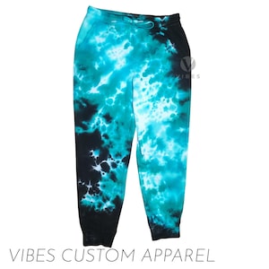 Op de afbeelding: Tie-dye joggingbroek in tinten van teal, turquoise en zwart. De broek heeft een tailleband met trekkoord en geribbelde enkels. De tekst "VIBES CUSTOM APPAREL" staat onderaan de afbeelding.