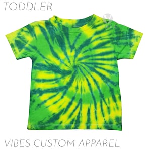 Może przedstawiać: Koszulka dla malucha w stylu tie-dye w odcieniach zieleni i żółci, ze spiralnym wzorem. Koszulka ma krótkie rękawy i okrągły dekolt. Nad koszulką znajduje się napis "TODDLER", a pod nią "VIBES CUSTOM APPAREL".