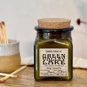 May include: A dark green glass jar candle with a cork lid. The label reads "Juniper Candle Co. Green Valley Lake Pine | Spruce | Honeysuckle Soy Candle Collection: Classic Burn Time: 45 Hours 7.7 oz / 220 g Green Valley Lake, CA"