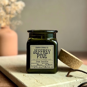May include: A dark green glass jar candle with a cork lid, labeled "JEFFREY PINE" with scent notes. The candle sits on a light-colored rectangular stone. A terracotta vase with dried flowers is in the background.