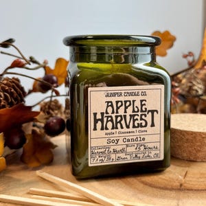 May include: A square, dark green glass jar soy candle with a cream-colored label reading Juniper Candle Co. Apple Harvest, Apple | Cinnamon | Clove, Soy Candle, Harvest to Hearth, 45 Hours, Net Weight 7.4 oz / 210 g, Green Valley Lake, CA.