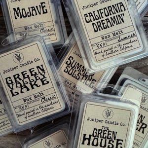 May include: Assortment of wax melts in clear plastic containers with labels featuring different scents and names, including "Mojave", "California Dreamin'", "Green Valley Lake", "Summer Solstice", and "The Green House". Each label has a handwritten font and a small illustration of a juniper branch.