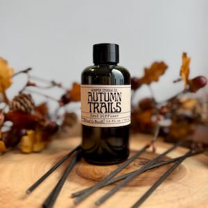 May include: A dark amber glass reed diffuser bottle with a black cap and a cream-colored label reading Juniper Candle Co. Autumn Trails Reed Diffuser 3.6 fl. oz. / 106 ml. Several thin black reeds are arranged around the base of the bottle.