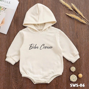 May include: Cream-colored baby romper with a hood and long sleeves. The text "Boba Corner" is printed on the front. The romper has snap closures at the bottom. The product code "SWS-06" is printed at the bottom.
