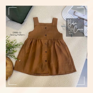 Puede incluir: Un vestido de lino marrón con botones en la parte delantera y tirantes. El vestido tiene una falda fruncida y está hecho con amor. El texto "DRBB-03 - Sewing Pattern -" y "Boba Corner Made with love" es visible en la imagen.
