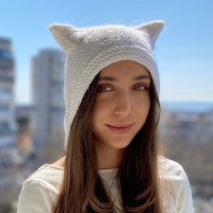 May include: A white knitted hat with cat ears. The hat has a hood and is made of a soft, fluffy yarn.