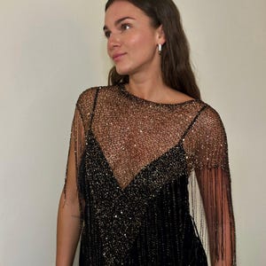 May include: A black dress with a beaded overlay featuring a V-neck design and fringe detailing. The overlay is a sheer, net-like fabric with gold-colored beads. The dress has a simple, elegant silhouette.