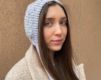 May include: A grey knitted hat with long ties. The hat is pulled down over the person's head and the ties hang down the back of their neck.