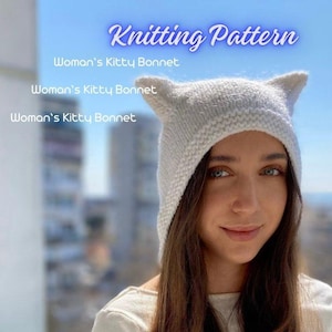 Könnte beinhalten: Weiße Strickmütze mit Katzenohren. Die Mütze hat eine spitze Spitze und einen umklappbaren Rand. Die Mütze wird von einer Person mit langen braunen Haaren getragen.