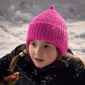 Op de afbeelding: Een felroze gebreide muts met een kleine pompon bovenop. De muts wordt gedragen door een persoon die een zwarte jas draagt, met een besneeuwde achtergrond. De muts lijkt gemaakt van dik, warm garen.
