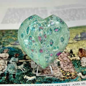 May include: A heart-shaped polished stone featuring a light green base color with scattered, vibrant magenta-pink inclusions throughout the surface. The stone is displayed on a small, clear plastic stand, showcasing its smooth, rounded texture.
