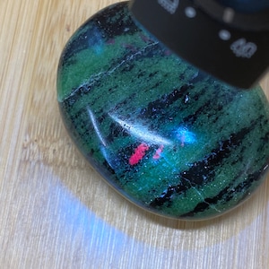May include: Close-up of a green and black gemstone with red inclusions under a UV light. The gemstone is smooth and polished.