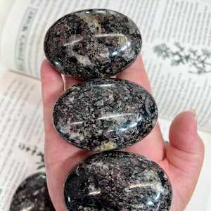 May include: Three polished oval stones with a speckled texture featuring a mix of dark black, grey, and deep red mineral inclusions. The stones have a smooth, glossy finish and are arranged in a vertical stack, with a fourth stone visible below.