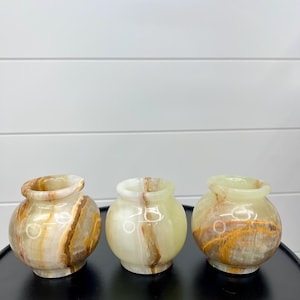 May include: Three small, round onyx vases with a polished finish. The vases have a light green and tan color scheme with natural veining patterns. The vases are displayed on a black surface against a white background.