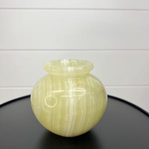 May include: A small, round, light green onyx vase with a narrow neck. The vase has a smooth, polished surface with vertical white veining. It sits on a black surface against a white background.