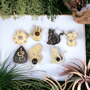 May include: Seven wooden ornaments with different designs, including a hand, a cat, a mushroom, a snake, and a sun and moon. Each ornament has a small hole in the center and is decorated with gold paint and intricate details. The ornaments are displayed on a white surface.