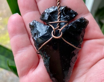 Pode incluir: Um pingente de ponta de flecha de obsidiana preta envolto em fio de cobre. A ponta de flecha é de um preto brilhante escuro e tem uma ponta afiada. O fio de cobre é enrolado em torno da ponta de flecha em um padrão decorativo.
