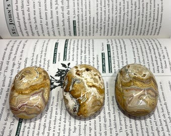 May include: Three oval-shaped polished stones with intricate banding in shades of brown, cream, white, and translucent gray. The stones are arranged in a horizontal row on top of an open book with printed text about St. John's Wort and sage.