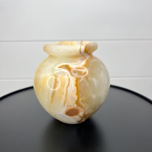 May include: A small, round onyx vase with a wide opening. The vase has a light beige base color with streaks of brown and white. The vase is approximately 4 inches tall.