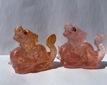 May include: Two translucent pink dragon figurines with gold accents. One dragon has a red eye, the other has a blue eye. The dragons are in a seated position with their tails curled.