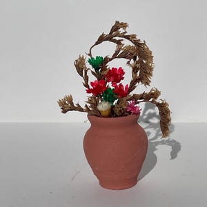 May include: A small, terracotta-colored ceramic vase filled with dried flowers. The arrangement includes red, green, and pink blooms, with dried brown stems and leaves. The vase is set against a white background.